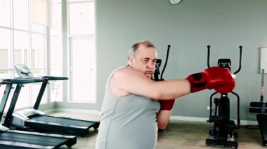 Kırmızı boks eldiveni giyen, spor salonuna yumruk atan kendinden emin, kilolu bir son sınıf öğrencisi. Sağlıklı ve kararlı bir yaşam tarzının parçası olarak boks yoluyla aktif yaşlanma, zindelik ve gücü teşvik eder..