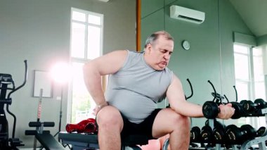 Spor salonunda biseps kıvrımlı otururken kilolu bir adam halterini kaldırıyor. Direniş eğitimi ve kişisel fitness hedefleri yoluyla güç, adanmışlık ve sağlıklı yaşlanma vurgulanıyor.