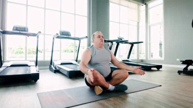 Parlak bir spor salonunda koşu bandı kullanmaya hazırlanan kilolu yaşlı bir adam sağlığını ve sağlıklı bir yaşam tarzını destekliyor. Fitness, sağlıklı yaşlanma ve kişisel refahı geliştirmeye odaklanmış..