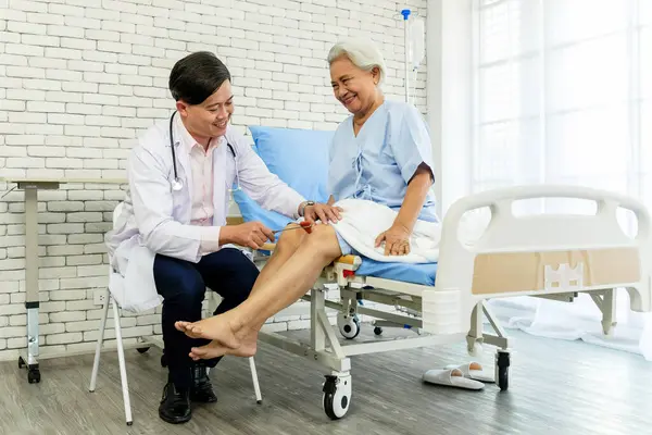 Bir doktor hastane kontrolü sırasında yaşlı bir kadının dizine refleks testi uygular. Sahne şefkatli bir bakım, tıbbi uzmanlık ve kıdemli sağlık ve sağlığın önemini gösteriyor.