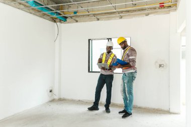 Miğfer ve güvenlik yelekleri giyen iki inşaat işçisi, görülebilir tavan boruları ve beton döşemeleri olan bitmemiş bir odada dijital tablet ve pano kullanarak bina planlarını tartışıyor..