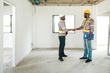 Güvenlik teçhizatı ve kask takan iki inşaat işçisi inşaat halindeki bir binanın içinde el sıkışıyor, takım çalışmasını, emlak gelişimini ve konut sektöründeki başarıyı simgeliyor..