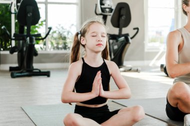 Yoga yapan genç bir kız gözleri kapalı ve elleri namaste pozisyonunda parlak bir spor salonunda bir kadının yanında duruyor. Sağlığı, sağlıklı yaşam tarzını, aile uygunluğunu ve huzurlu ev ortamlarını teşvik eder..