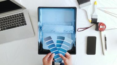 Woman selects blue color swatches while preparing for a DIY wall painting project. Home renovation tools, laptop, and roller tray reflect creativity, planning, and interior makeover inspiration.