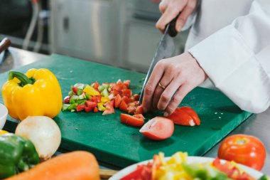 Beyaz üniformalı bir şef, yeşil bir kesme tahtasında taze domatesleri dilimliyor. Profesyonel bir mutfakta doğranmış dolmalık biberle. Yemek kursunda renkli ve sağlıklı bir yemek için malzemeler hazırlıyor..