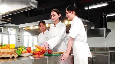 Aşçılık eğitmeni, modern bir ticari mutfakta, taze sebzelerle elle pişirme kursunda iki genç aşçıya rehberlik ediyor. Aşçılık okulu, mutfak sanatı ve yemek eğitimi için ideal..