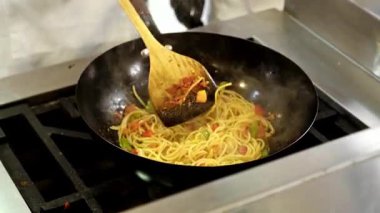 Fırında kızartılmış sebzeli spagetti pişiren bir tavanın yakın çekimi. Ahşap bir spatula, yemek hazırlama veya mutfak içeriği teması için ideal bir ticari mutfakta buharda erişte karıştırır..