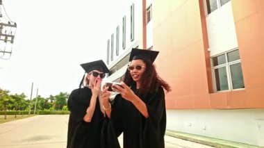 Siyah önlüklü ve şapkalı iki neşeli bayan açık havada neşeli bir selfie çekiyor. Kollarını kaldırıp gülümseyerek modern bir üniversite binası önünde akademik başarıyı kutluyorlar..