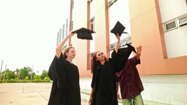Dışarıda kutlama yapan üç genç öğrenci, şapkalarını neşe ve gururla kaldırıyor. Mutlu grup, akademik başarılarını ve geleceklerini simgeleyen kampüs binası yakınlarında güven içinde yürüyor. Yavaş çekim 4k