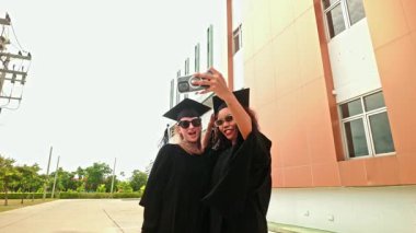 Siyah önlüklü ve şapkalı iki bayan açık havada neşeli bir selfie çekiyor. Parlak bir şekilde gülümserler. Mezuniyet günlerinde kutlama, başarı ve dostluk dolu bir an. Yavaş çekim 4k video.