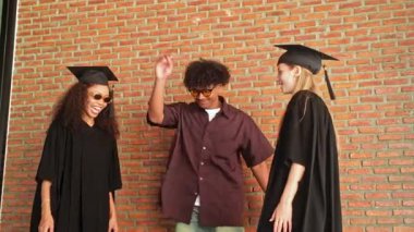 Mutlu mezunlar ve bir arkadaş, duvara karşı beşlik çakarak kutluyorlar. Neşeli an, akademik bir dönüm noktasını tamamladıktan sonra gurur, başarı ve beraberliği yakalar. 4k gerçek zamanlı video.