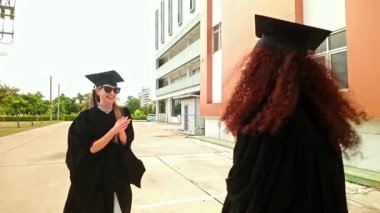 Şapka ve cüppeli iki mutlu mezun dışarıda selfie çekiyorlar. Gülümseyip kollarını kaldırarak, parlak bir mezuniyet gününde başarı ve dostluk anını yakalıyorlar. Gerçek zamanlı 4k video