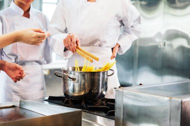 Beyaz şef üniformaları içinde yemek pişirme öğrencileri fırının üstünde paslanmaz çelik bir tencerede makarna hazırlıyorlar. Ticari bir mutfakta elle yapılan bir aşçılık dersi sırasında temel yemek hazırlama becerilerini öğreniyorlar..