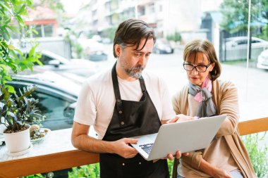 Kafe sahibi ve işletme danışmanı akıllı telefonlar kullanarak pazarlama veya finansmanı tartışıyor. Her ikisi de gülümsüyor ve meşgul, takım çalışmasını, stratejiyi ve modern küçük iş ortaklığını sembolize ediyor..