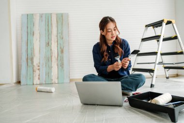 Genç bir kadın dizüstü bilgisayarında araştırma yaparken elinde boya fırçası tutuyordu. Aydınlık bir odada aletler, merdiven ve ahşap paneller ile çevrili, ev geliştirme planlama odaklı.