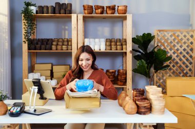 Genç bir kadın, küçük bir iş yerinde mavi baloncuklu ambalaja sarılı kırılgan bir eşyayı açarken gülümsüyor. Ürün güvenliği, ambalaj, e-ticaret ve envanter kalite kontrolü kavramı.