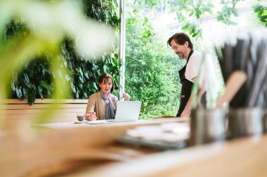 Cafe sahibi yeşil temalı bir kafede finans danışmanıyla görüşüyor. Önlüklü adam dinlerken iş elbiseli kadın bilgisayardaki belgeleri inceliyor. Doğal ışık ile kentsel eko ayarı.