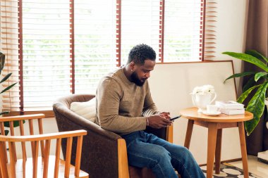 Rahat bir oturma odasında oturan, akıllı telefonuna odaklanmış rahat bir adam. Parlak doğal ışık, kapalı alan bitkileri, kahve fincanı ve kitaplar sıcak, otantik bir yaşam tarzı ve ev konforu ortamı yaratır..