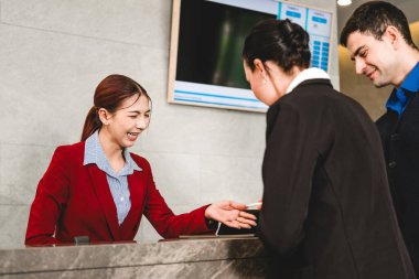 Kırmızı ceketli gülümseyen otel resepsiyonisti resepsiyondaki müşterilere sıcak ve verimli giriş hizmeti veren bir anahtar kartı veriyor. Hoş bir seyahat ortamında dostça bir konukseverlik..