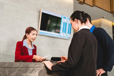 Kırmızı ceketli bir otel resepsiyonisti kayıt sırasında misafirlere yardım ediyor, belgeleri teslim ediyor. Modern bir seyahat ve konukseverlik ortamında dostane ve profesyonel bir hizmet etkileşimi.