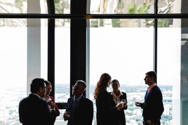 Çeşitli profesyonellerden oluşan bir grup, yüksek katlı bir ofisin yüksek pencerelerinin yanında gelişigüzel konuşuyor. İşten sonra şehir görüşleriyle dinlenme sohbeti, ağ oluşturmayı ve takım bağlantısını teşvik eder.