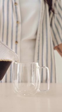 Taze kahve sürahisinden, arka planda çizgili gömlekli bir kişinin olduğu, parlak bir mutfak tezgahının üzerindeki çift duvarlı bardağa dökülüyor..