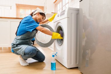 Eldivenli biri, parlak bir mutfakta çamaşır makinesini temizliyor, varili bezle silmek için diz çöküyor, yere bir şişe temizleyici sıkıyor..