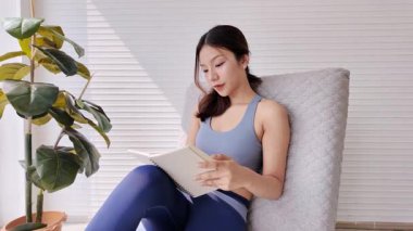 Evde tablet ve stil kullanan atletik giyimli genç bir kadın, bir ev bitkisi ile bir pencerenin yanında oturuyor, işe ve rahatlamaya odaklanıyor.