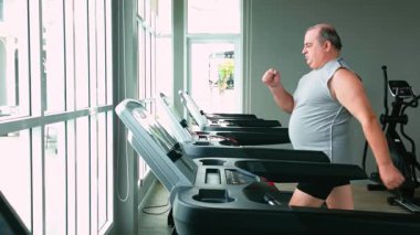 Parlak bir spor salonunda koşu bandında koşan, makineler ve büyük pencerelerle dolu, spor yaparken müzik dinleyen ve spor yaparken vücut ve kardiyo eğitimine odaklanan bir adam..