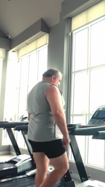 Büyük pencereli, spor aletleri ve kulaklıklar takmış, kardiyo egzersizi ve sağlıklı, aktif bir yaşam tarzına odaklanmış, koşu bandında yürüyen adam..