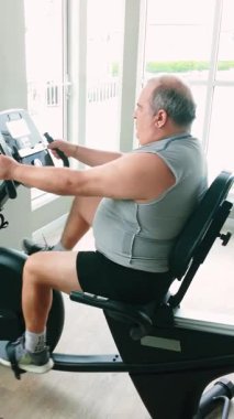 Parlak bir spor salonunda yaslanmış bisiklet üzerinde egzersiz yapan bir adam, kulaklıklar ve konsolda takip ölçümleri kullanarak; kapalı alanda kardiyo çalışması ve sağlıklı yaşam tarzı konsepti.