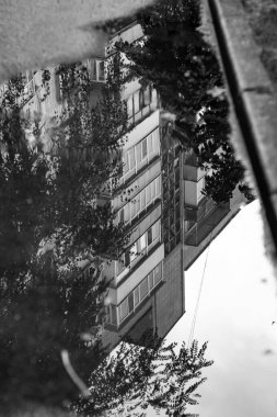 Tek renkli dikey fotoğraf, suya yansıyan balkonlu konut binası, yağmurdan sonra birikinti..