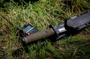 Taktiksel bir askeri bıçak, telsizin yanındaki kuru çimlerin üzerinde uzanıyor. Bıçak sağlam bir kılıfın içinde ve telsizin anten ve görünür kontrol düğmeleri var..