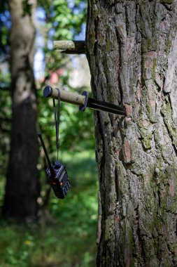 Sapında bir telsiz cihazı asılı duruyor. Radyo portatif görünüyor, düğmeleri ve yukarı bakan bir anteni var.. 