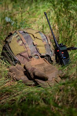 Kamuflaj desenli askeri miğfer ve yeşil çimlerin üzerinde duran siyah bir telsiz. Miğferde ağ ve ayarlanabilir kayışlar var. Telsiz görünür bir anten ile dijital bir ekran görüntüler. 