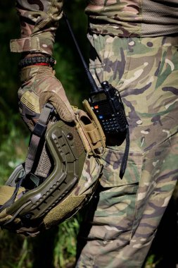 Taktik eldivenli bir asker elinde telsiz, askeri mühimmat, kamuflaj ve askeri işler olan bir saldırı kaskı tutuyor. Askeri üniforma.