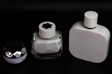 Siyah arka planda üç kozmetik konteynır var. Metal kapaklı küçük yuvarlak bir kavanoz, geniş beyaz kapaklı dikdörtgen bir şişe..