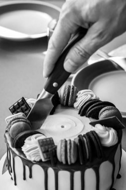 Pürüzsüz, hafif şekerli yuvarlak pasta ve kenarlarından siyah çikolata damlıyor. Bıçaklı pasta kesen el. Arka planda mavi jantlı tabaklar var, tatlı servisi..