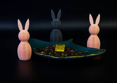 Üç stilize edilmiş tavşancık figürü, iki pembe ve bir gri, yaprak şeklindeki yaprak şeklindeki bir çanak, bol yapraklı çay, gül yaprakları, ve altın bir küp, hepsi koyu siyah bir zemin üzerinde..