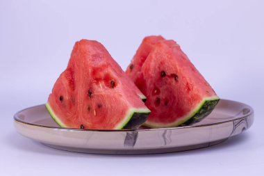 Dört dilim karpuz parlak kırmızı et ve siyah çekirdekli metal kenarlı yuvarlak bir tabakta yatıyor. Karpuz dilimlerinin beyaz kenarlı yeşil bir derisi var, tabakta düzgünce istiflenmiş..