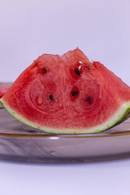 Yuvarlak bir tabakta parlak kırmızı merkezli bir karpuz parçası yatıyor. Karpuzun arka planında birkaç dilimle dilimin yüzeyine saçılmış yeşil deri ve tohumları vardır..