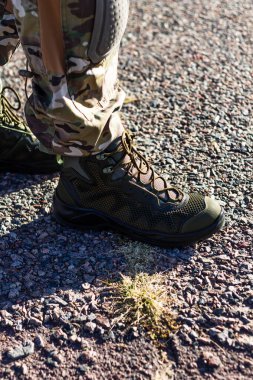 Bir askerin yeşil taktik botunun küçük bir çimen parçasıyla güneşli çakılların üzerinde duruşu. Çağdaş, dayanıklı askeri ayakkabı ve kamuflaj takımlarına odaklanın..