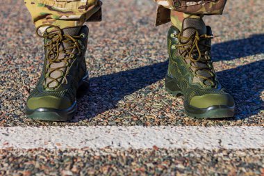 Beyaz bir çizmenin arkasında asfalt üzerinde duran yeşil taktik botlar ve kamuflaj pantolonları. Parlak güneş ışığı altında modern, dayanıklı askeri ayakkabılara odaklanın..