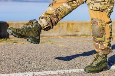 Tam bir askeri kamuflaj pantolonu giymiş, dizlikleri ve sağlam zeytin yeşili taktik çizmeleri olan, asfalt yolda yürüyen bir adamın dinamik bir fotoğrafı..
