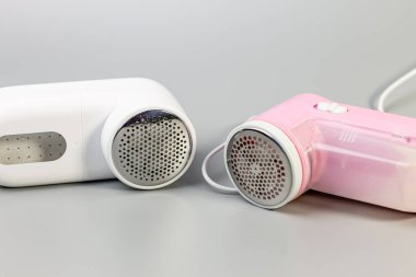 Tarafsız gri bir arkaplanda iki elektrikli kumaş traş makinesinin (beyaz ve pembe) yakın çekimi. Giysilerden iplik ve hap çıkarmak için modern ve temel araçlar.