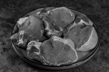 Görünür yağlı ve dokulu yüzeyli dört adet çiğ ve beyaz domuz bifteği, zenginleştirilmeye hazır, ahşap bir arka planda, yuvarlak bir tabakta sunuluyor..