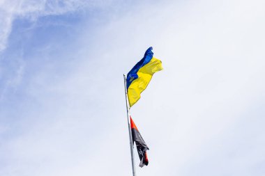 İki bayrak (Ukrayna ve kırmızı-siyah bayraklar) parlak, dramatik bir gri tonda gökyüzüne karşı bir direğin üzerinde dalgalanıyor ve ulusal ruhun ve dayanıklılığın tek renkli sembolünü oluşturuyor..