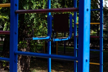 Canlı mavi metal çubuklar açık hava oyun alanı ya da sokak antrenman sahasının ön planına hükmediyor, arkada yumuşak odaklı yemyeşil ağaçlar dinamik bir renk ve derinlik patlaması yaratıyor..