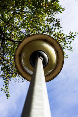 Aşırı düşük açılı klasik sarı sokak lambası manzarası ve açık mavi gökyüzüne uzanan sırık, yeşil ağaç yaprakları çerçeveli, doğa ve kentsel karşıtlık.