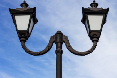 Çarpıcı, simetrik yakın plan, yumuşak bulutlu canlı mavi gökyüzüne karşı ikiz bir lamba direği. Süslü metal tasarımı klasik şehir zarafetini gösteriyor..